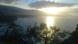 Paesaggio al tramonto con il sole che si riflette sulla superficie del mare, profili di rami in primo piano e montagne che sfumano all'orizzonte sotto un cielo nuvoloso.